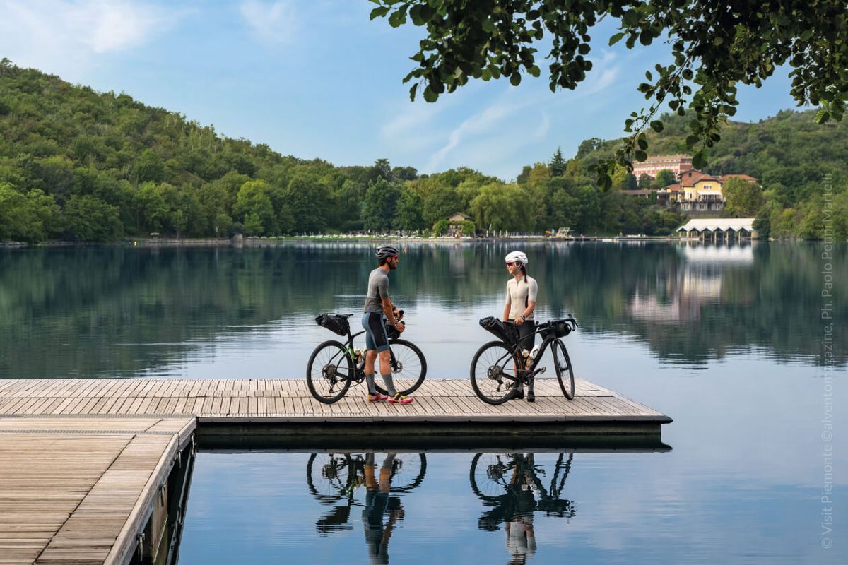 Due cicloturisti con bici da bikepacking sostano su un pontile di legno che si specchia in un lago circondato da colline boscose